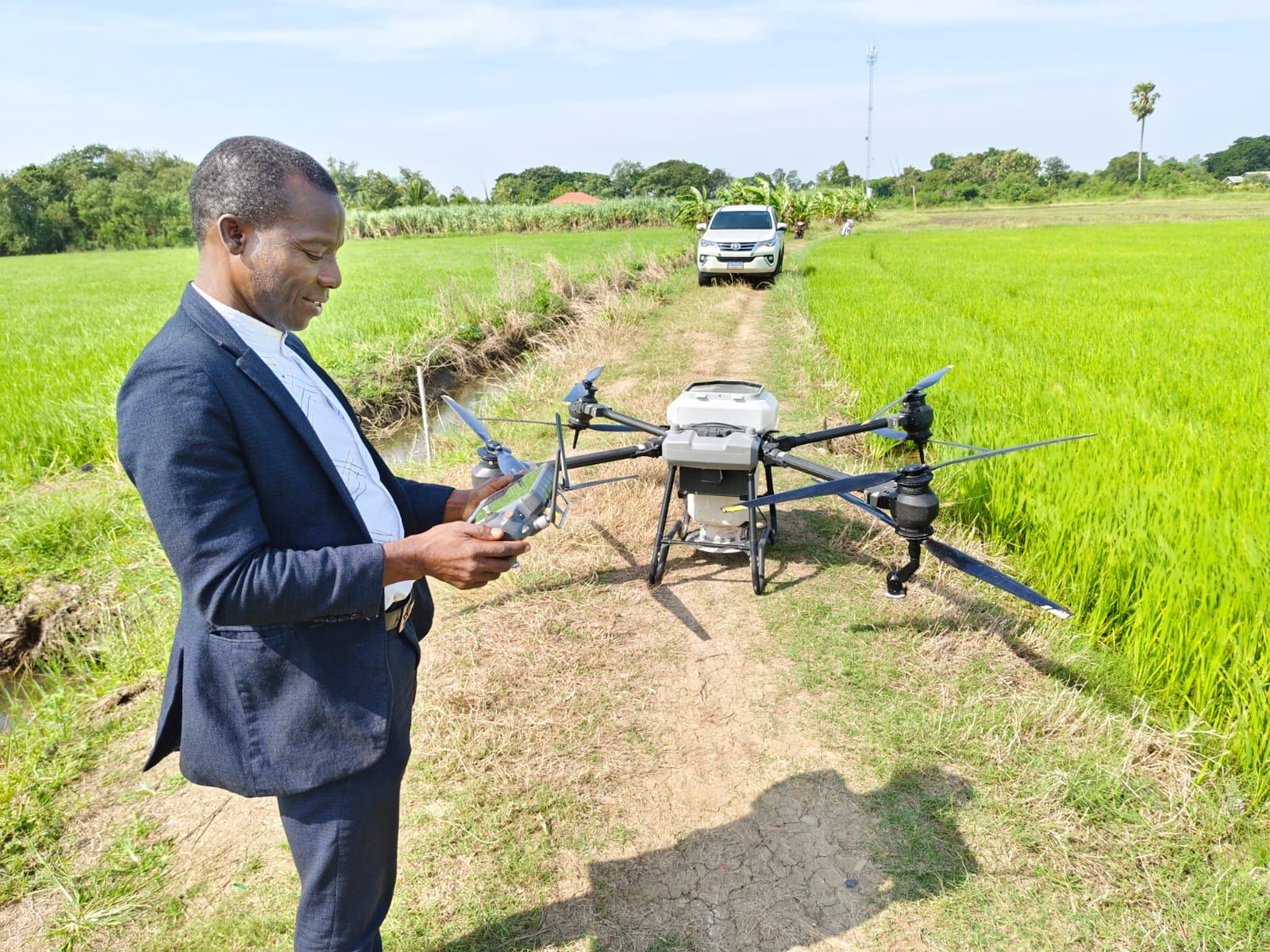 Programme lead holding a drone controller beside a spraying drone in a rice field